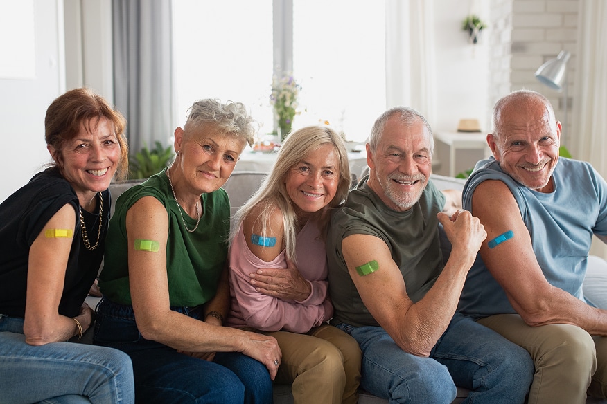 A person with gray hair and beard smiling with a bandaid on their arm